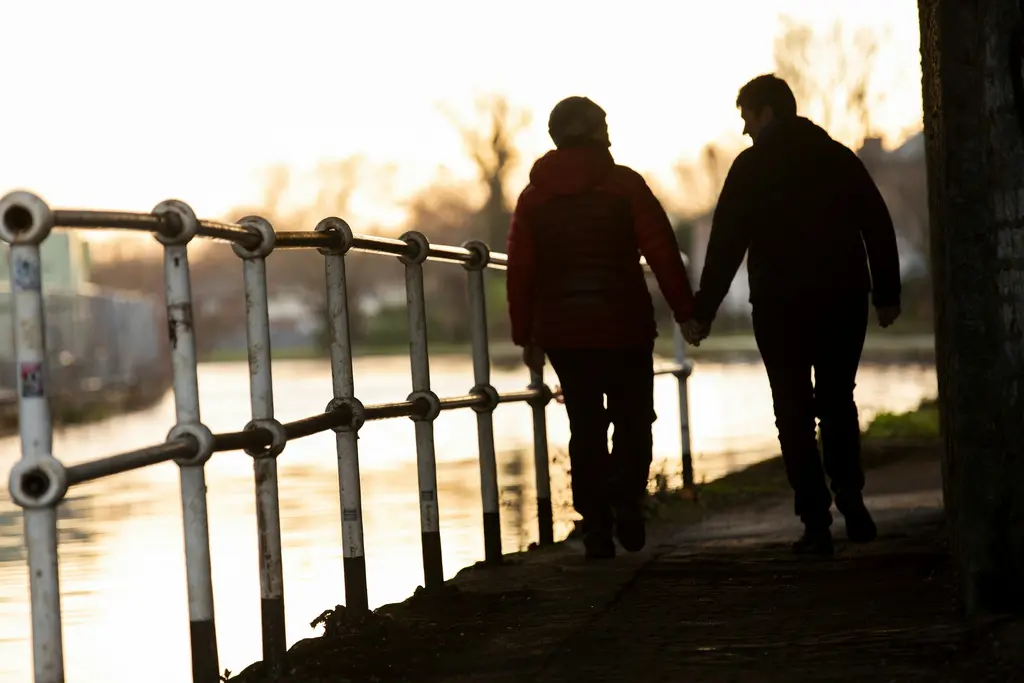 An older couple walk hand in hand along a riverside path in the evening light.