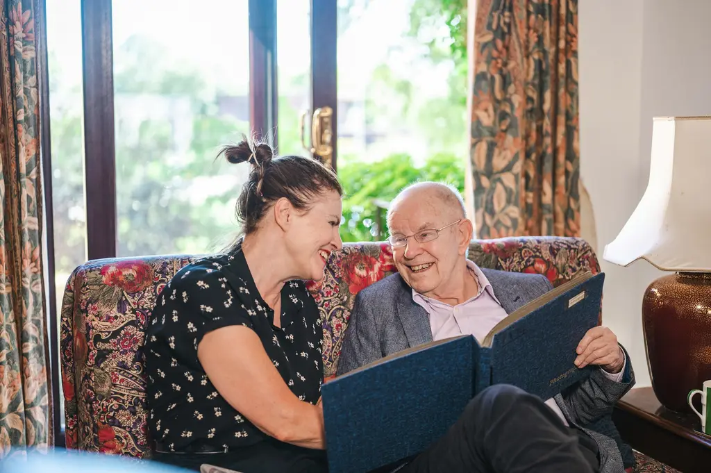 An older man and a younger woman laugh together on his sofa while looking through a photo album.