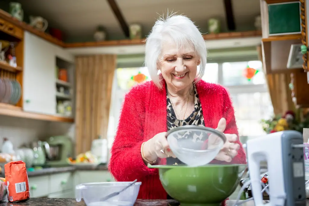 A woman with white hair smiles as she sieves flour into a bowl in her own kitchen.